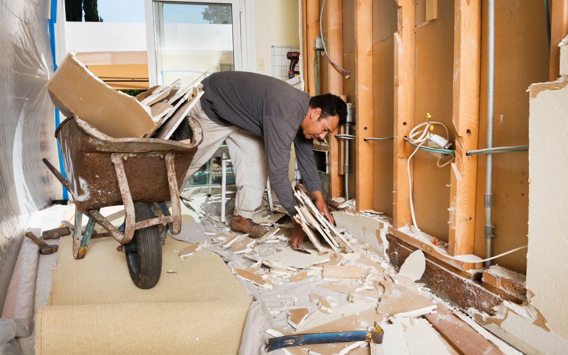Worker clearing demolition debris from a kitchen remodel, with exposed wall framing, visible plumbing and electrical lines, and a wheelbarrow filled with broken drywall