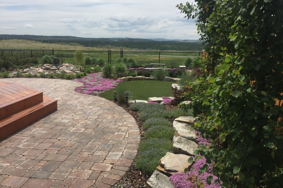 Curved interlocking paver walkway with colorful flowering garden beds, a stone retaining wall, and open lawn, a landscape construction project in Calgary