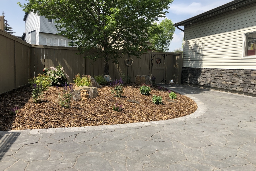 Backyard interlocking paver patio with a curved mulch garden bed, mature tree, and stone accent wall, landscape construction in Calgary by Western Elements Landscaping