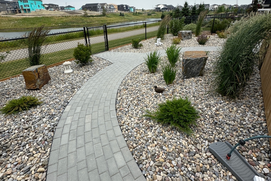 Curved backyard interlocking paver walkway through a wide river rock garden with ornamental grasses and boulder accents, a landscape in Calgary by Western Elements Landscaping