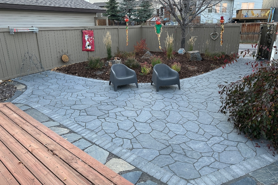Backyard flagstone-pattern interlocking paver patio with modern lounge chairs and a dark mulch garden bed, a landscape design and build in Calgary