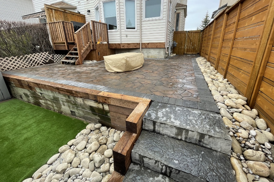 Raised flagstone-pattern interlocking paver patio with timber retaining walls, river rock border, and a covered fire pit, a landscape in Calgary by Western Elements Landscaping