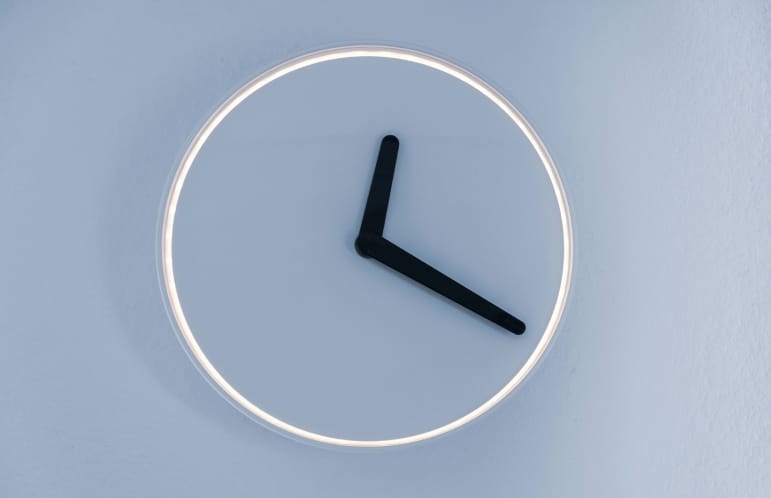 Minimalist wall clock with black hour and minute hands and a glowing circular frame on a light blue wall.