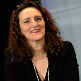 Smiling woman with curly brown hair wearing a black jacket and pearl necklace.