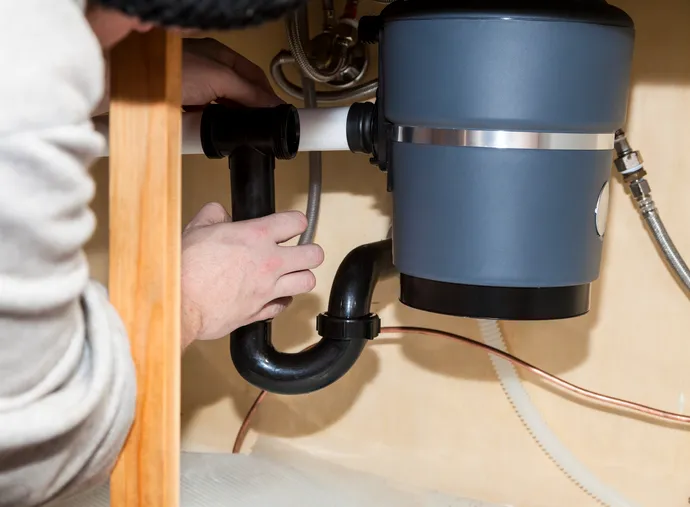 Close-up of a person connecting the black P-trap pipe to a gray garbage disposal unit under a kitchen sink.