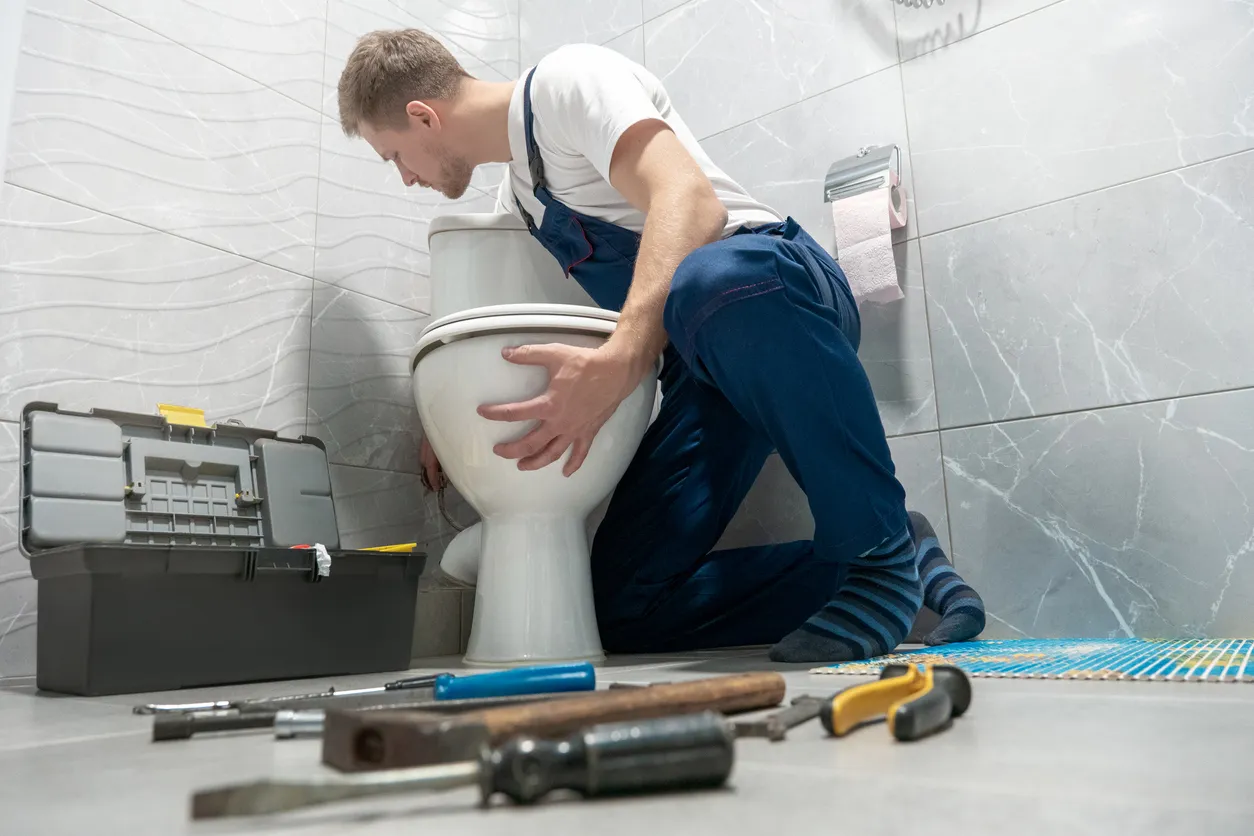 A plumber installs a new toilet bowl in a tiled bathroom with a toolbox and various tools scattered on the floor.