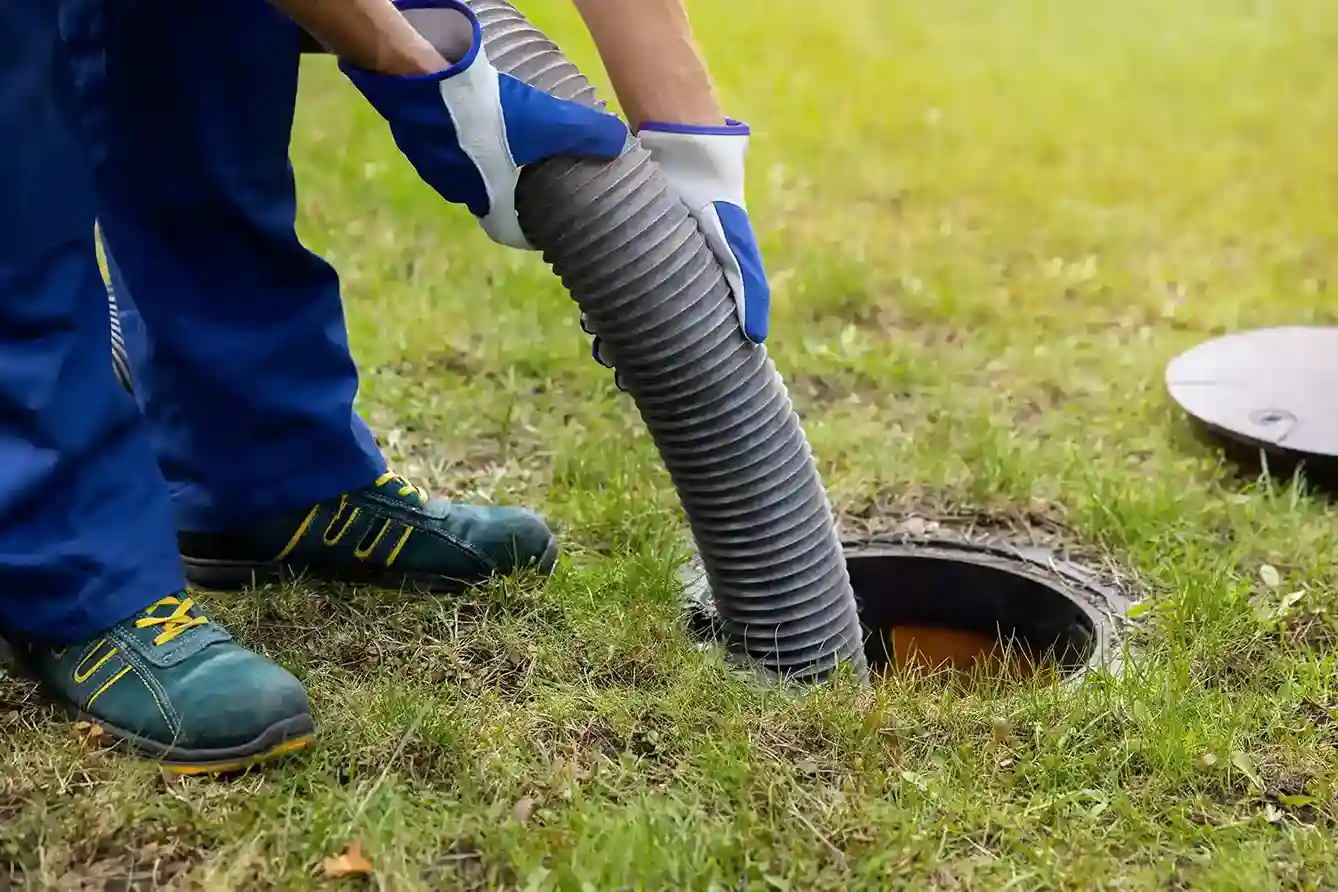 Plumber wearing gloves inserting a large septic pump hose into an outdoor manhole or sewer drain.