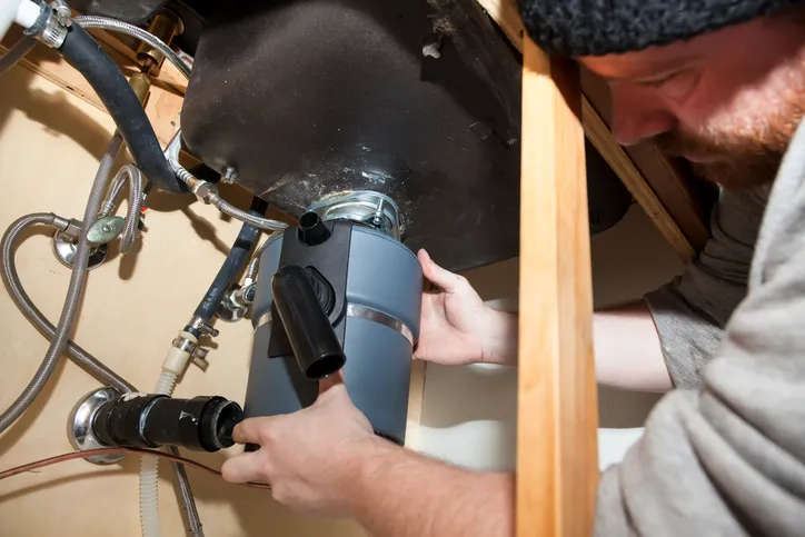 Man with a beard installing a gray garbage disposal under a sink, connecting the drain pipe.
