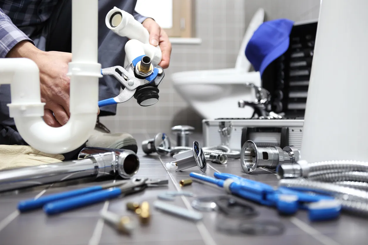 Close-up of a plumber connecting white PVC and chrome drain pipes with a wrench, surrounded by plumbing parts.