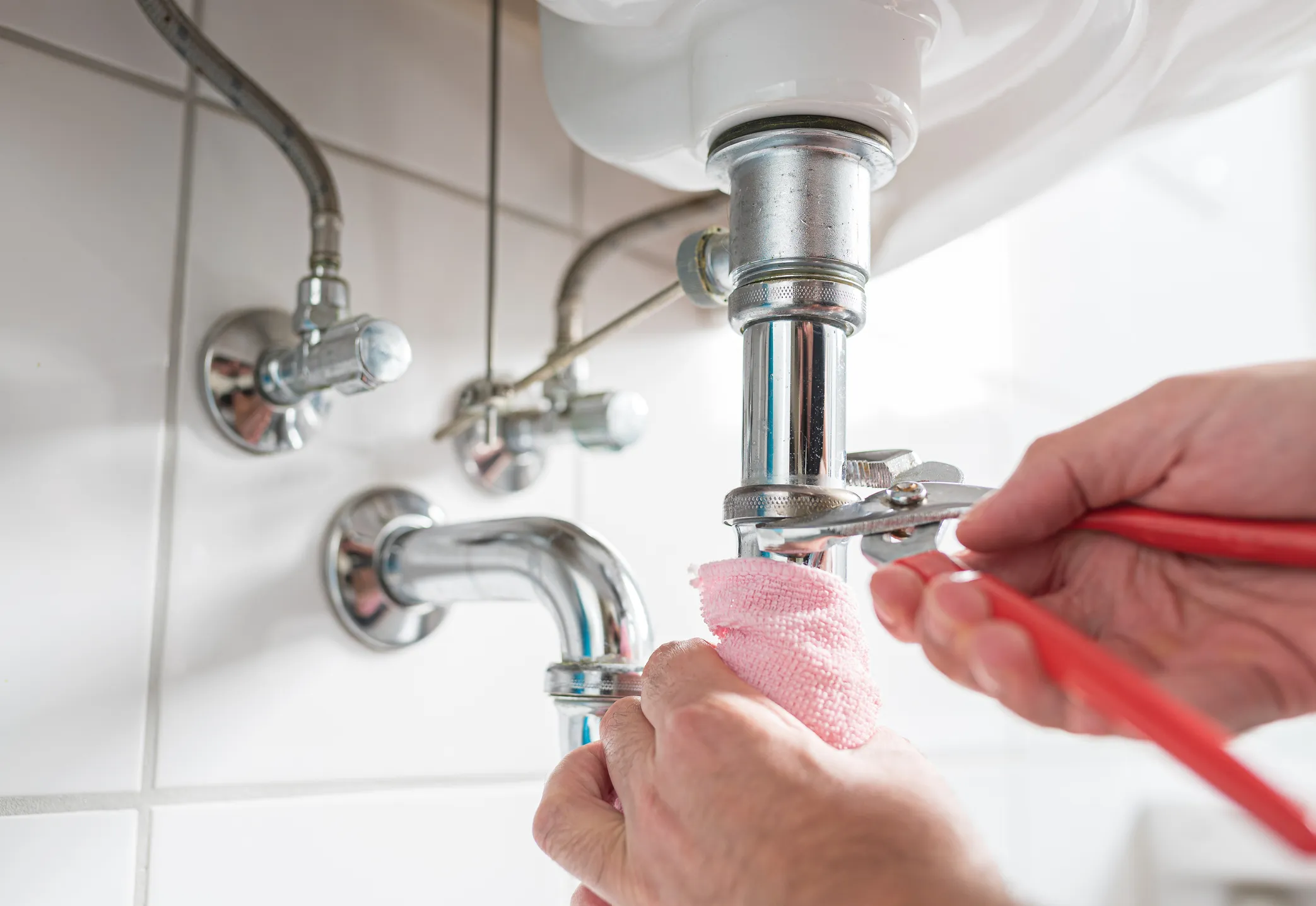 Close-up of hands using pliers to tighten a bathroom sink drain pipe with a pink cloth.