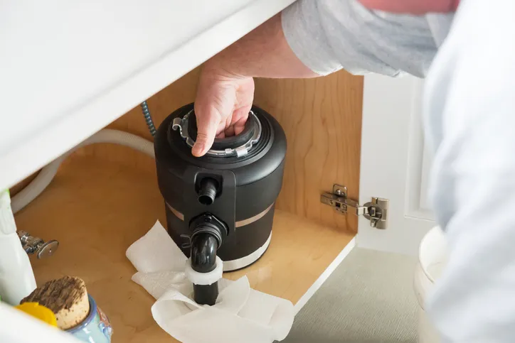 Close-up of a person installing a black garbage disposal unit under a kitchen sink, with paper towels on the cabinet base.