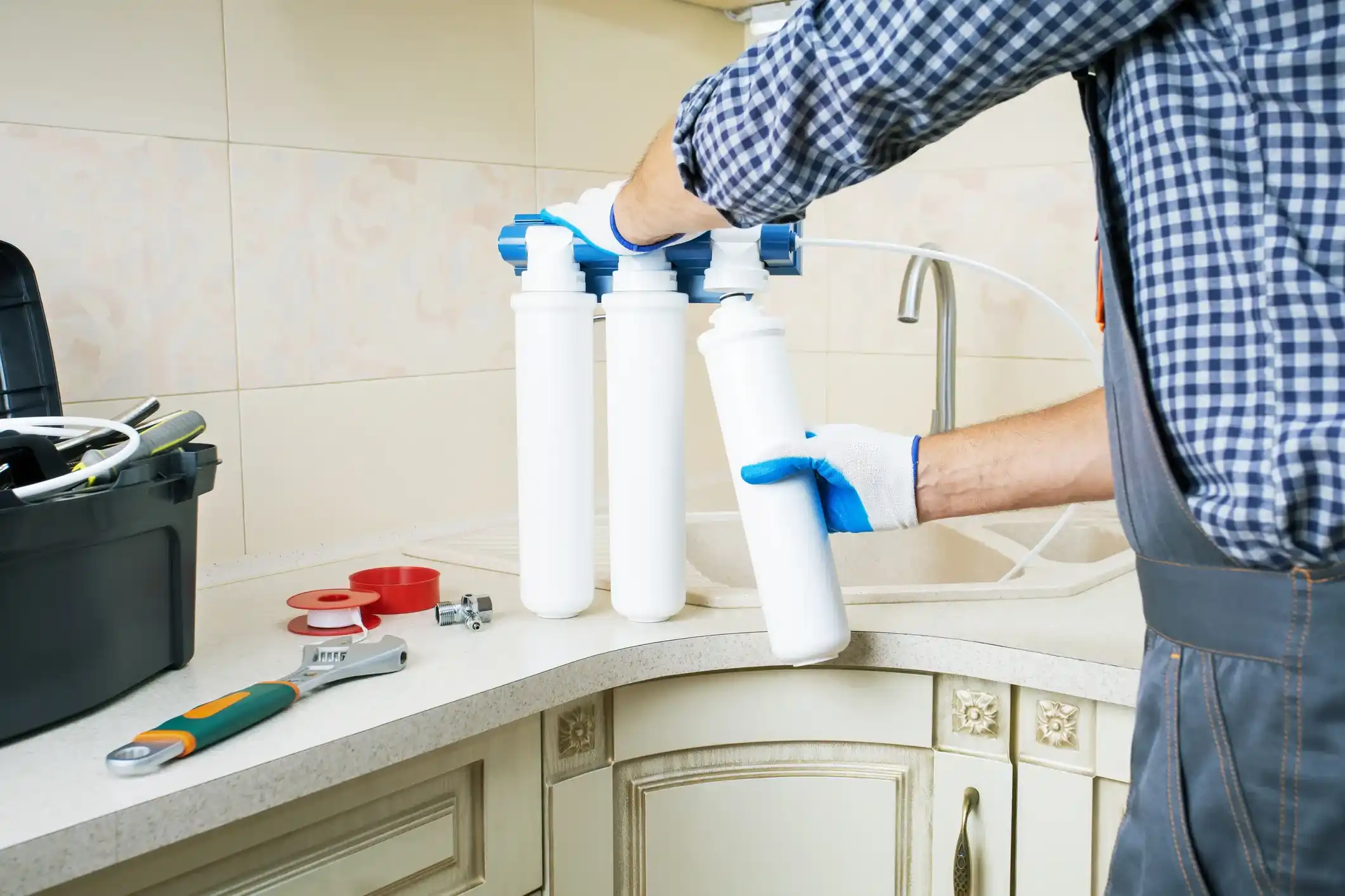 A plumber holds a white cylindrical water filter cartridge, preparing to install it into a multi-stage system on a kitchen counter.