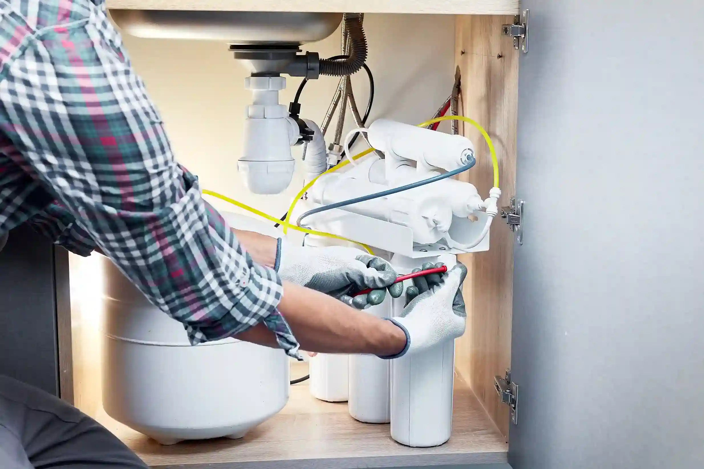 A person uses a screwdriver to perform maintenance on an under-sink water filtration system and its storage tank.