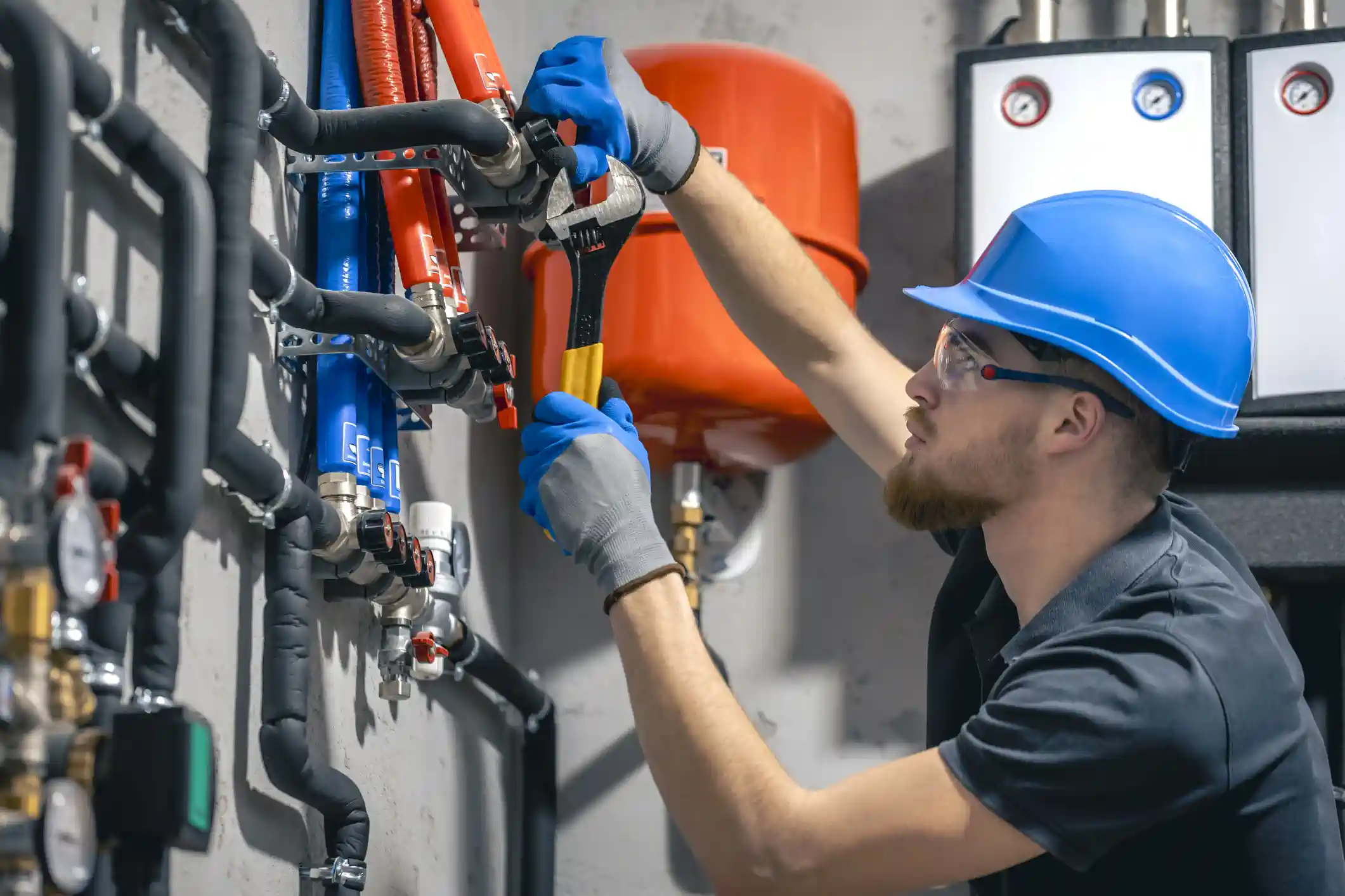  A male HVAC or plumbing technician is working on a heating distribution manifold in a commercial or mechanical room. He is wearing a blue hard hat, safety glasses, and grey and blue work gloves, and is using a wrench to tighten a fitting on the system. The manifold features numerous pipes, some insulated in black, with distinctive red and blue sections indicating flow lines. A large, spherical orange expansion tank is visible in the background, along with a white control panel. The overall impression is professional installation or maintenance of a complex heating system.