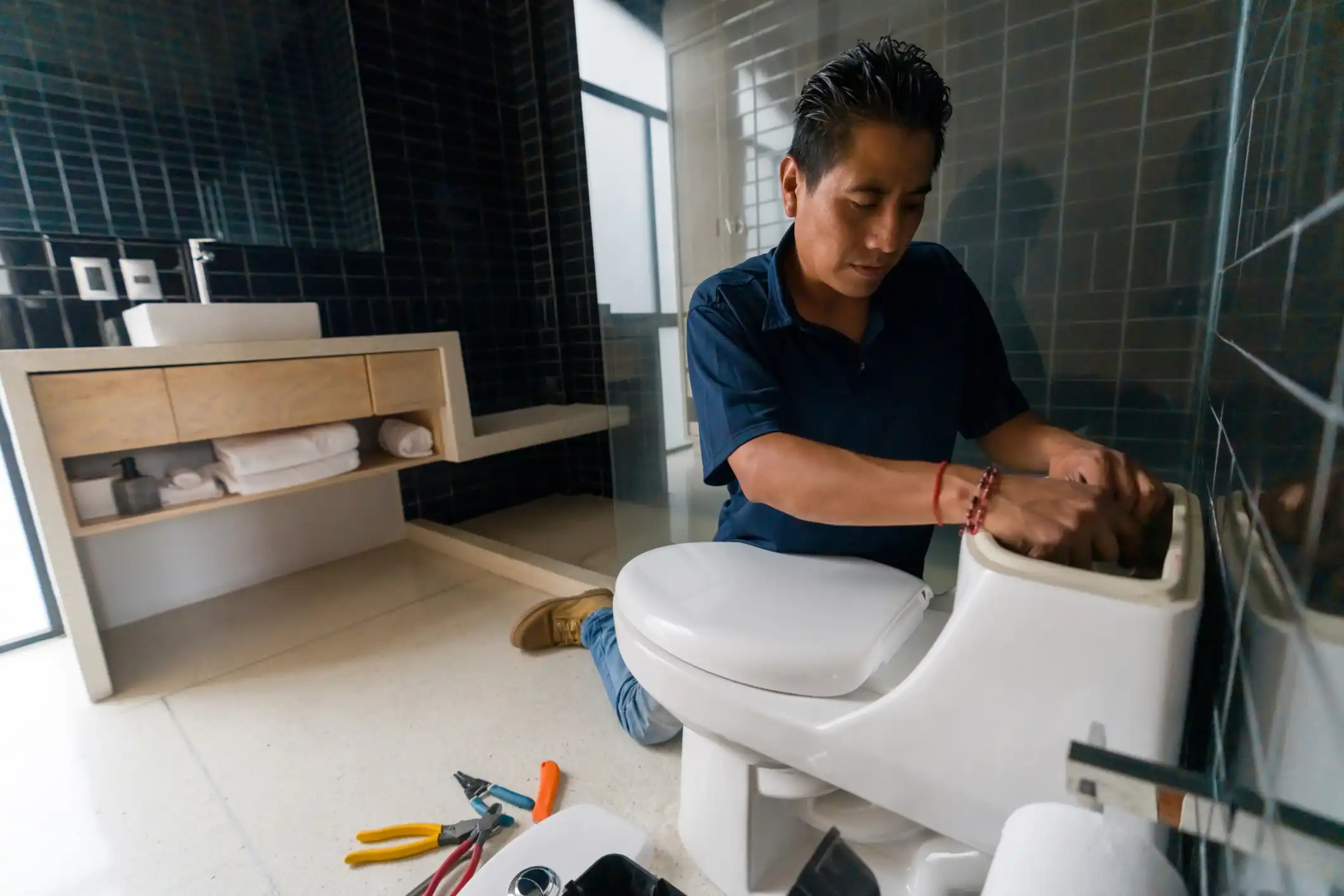 A man is kneeling on a tiled floor, repairing the inner components of a toilet tank.