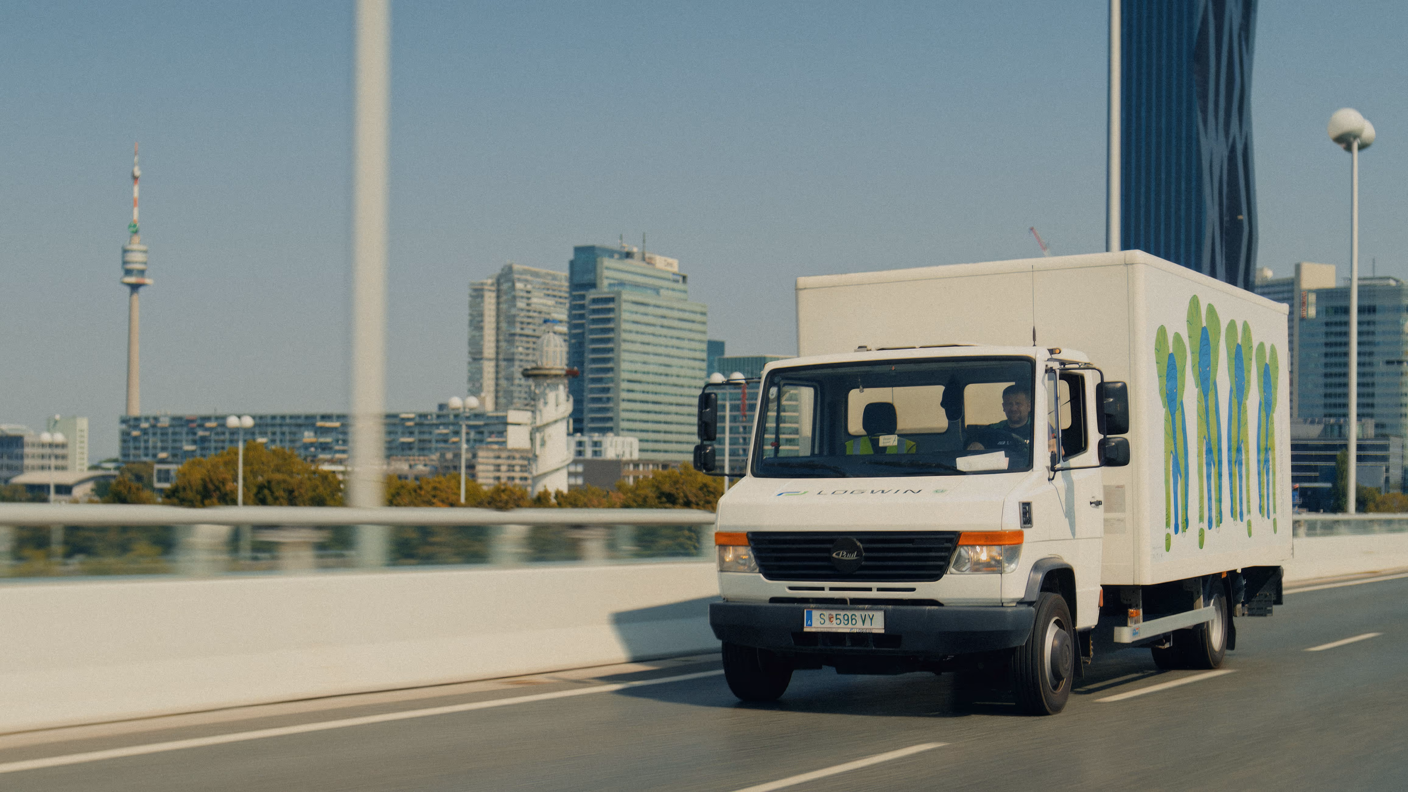 Weißer Logwin-Transporter mit grün-blauer Grafik fährt auf einer Stadtautobahn mit Hochhäusern im Hintergrund.