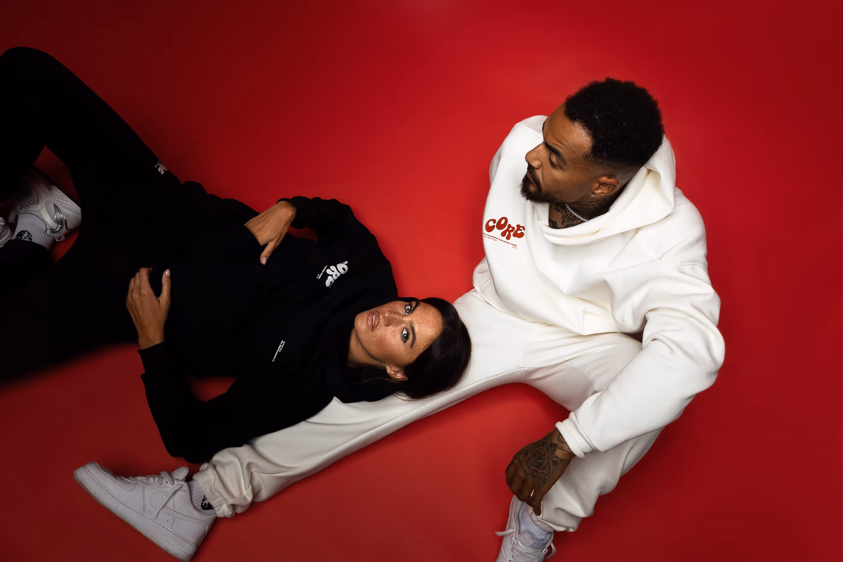 Ein Mann und eine Frau in lässiger Streetwear posieren auf rotem Boden, der Mann trägt weiße Kleidung, die Frau schwarze.
