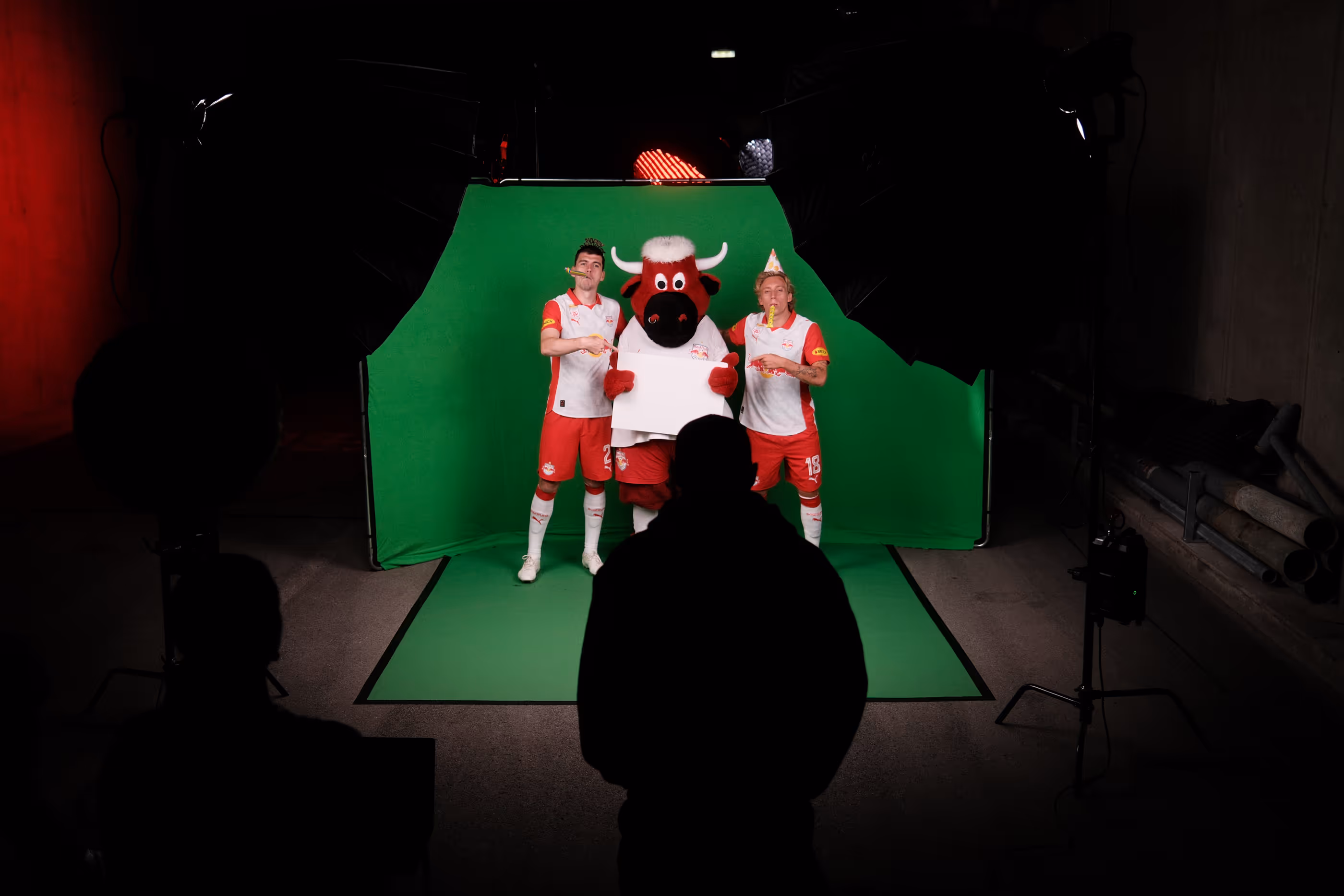 Zwei Fußballspieler und ein Maskottchen vor einem grünen Hintergrund mit Partyhüten und Tröten in einem Fotostudio.