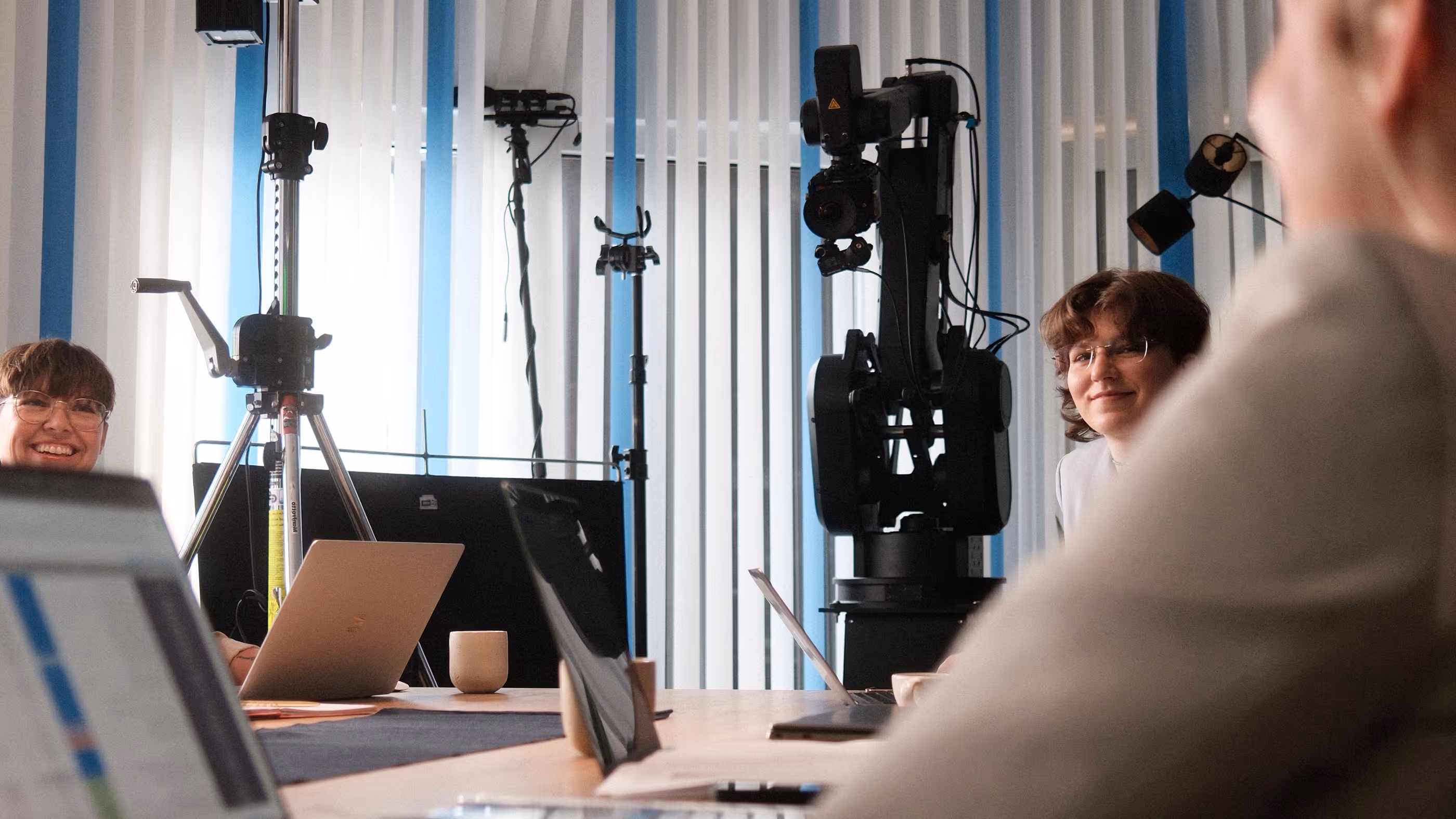 Drei Personen in einem gut beleuchteten Büro sitzen lächelnd an einem Tisch mit Laptops und einer großen Kamera oder einem Roboterarm im Hintergrund.