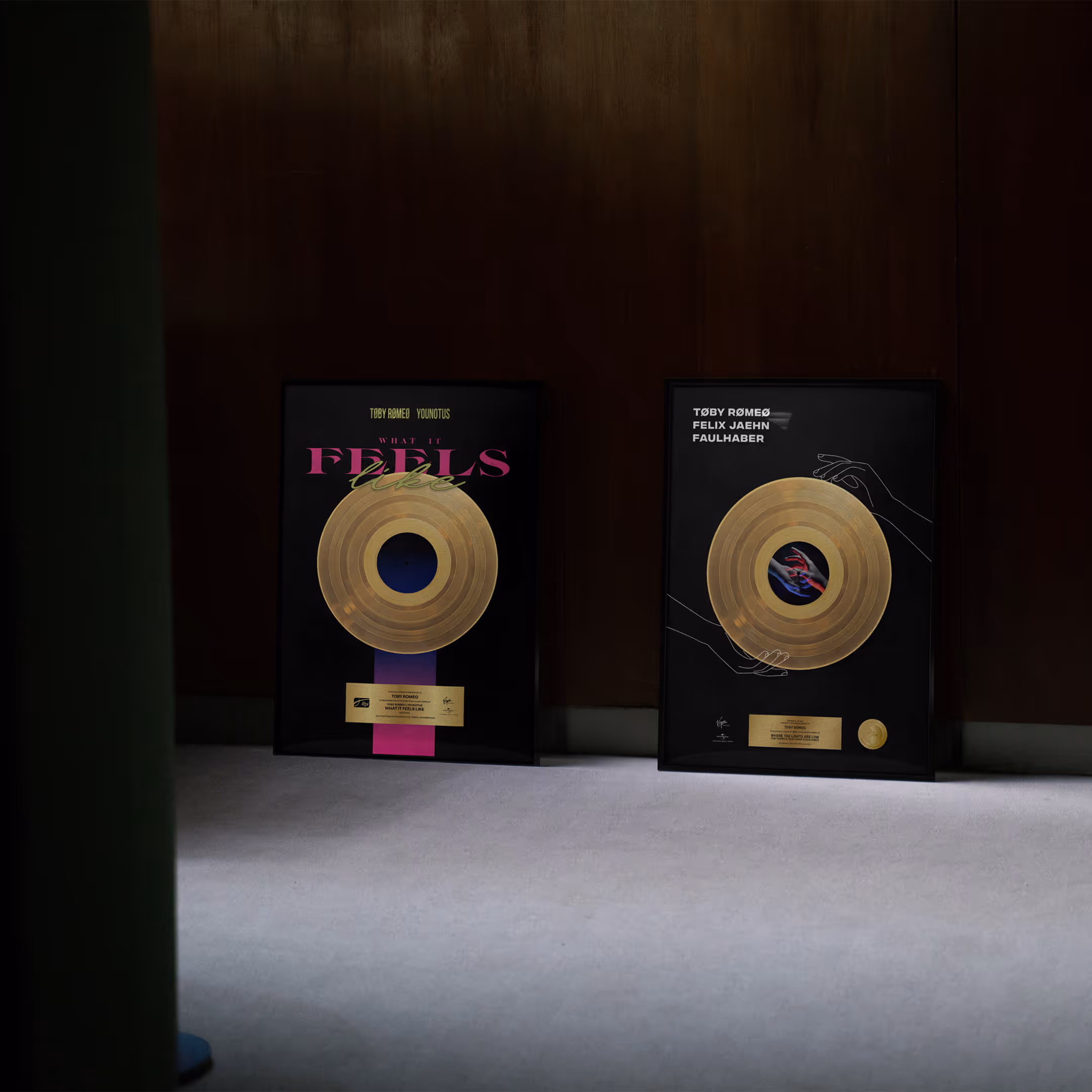 Zwei gerahmte goldene Schallplatten mit schwarzen Hintergründen lehnen vor einer dunklen Holzwand auf dem Boden.