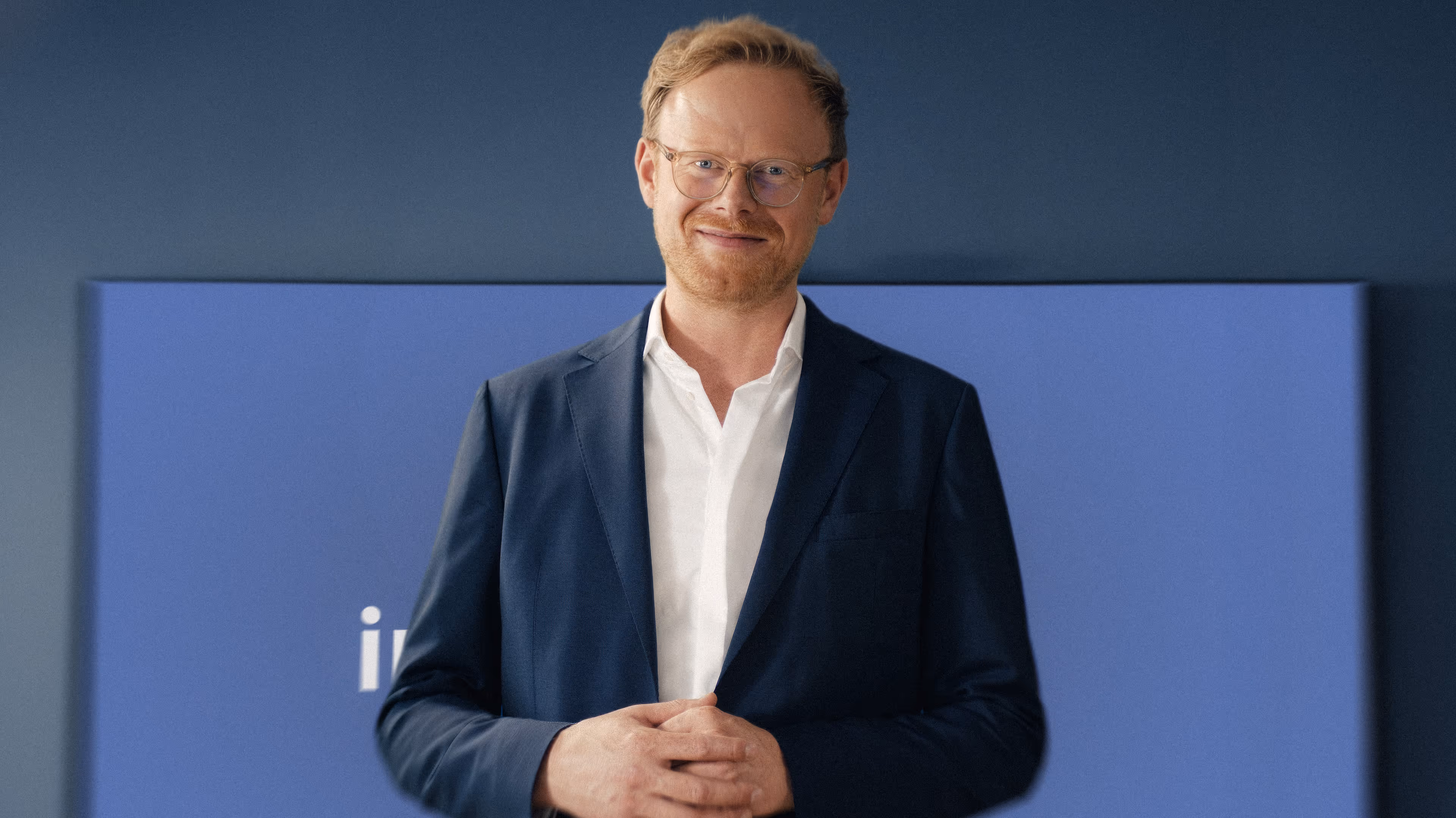 Lächelnder Mann mit Brille, blonden Haaren und Bart, gekleidet in einem weißen Hemd und blauem Sakko vor blauem Hintergrund.