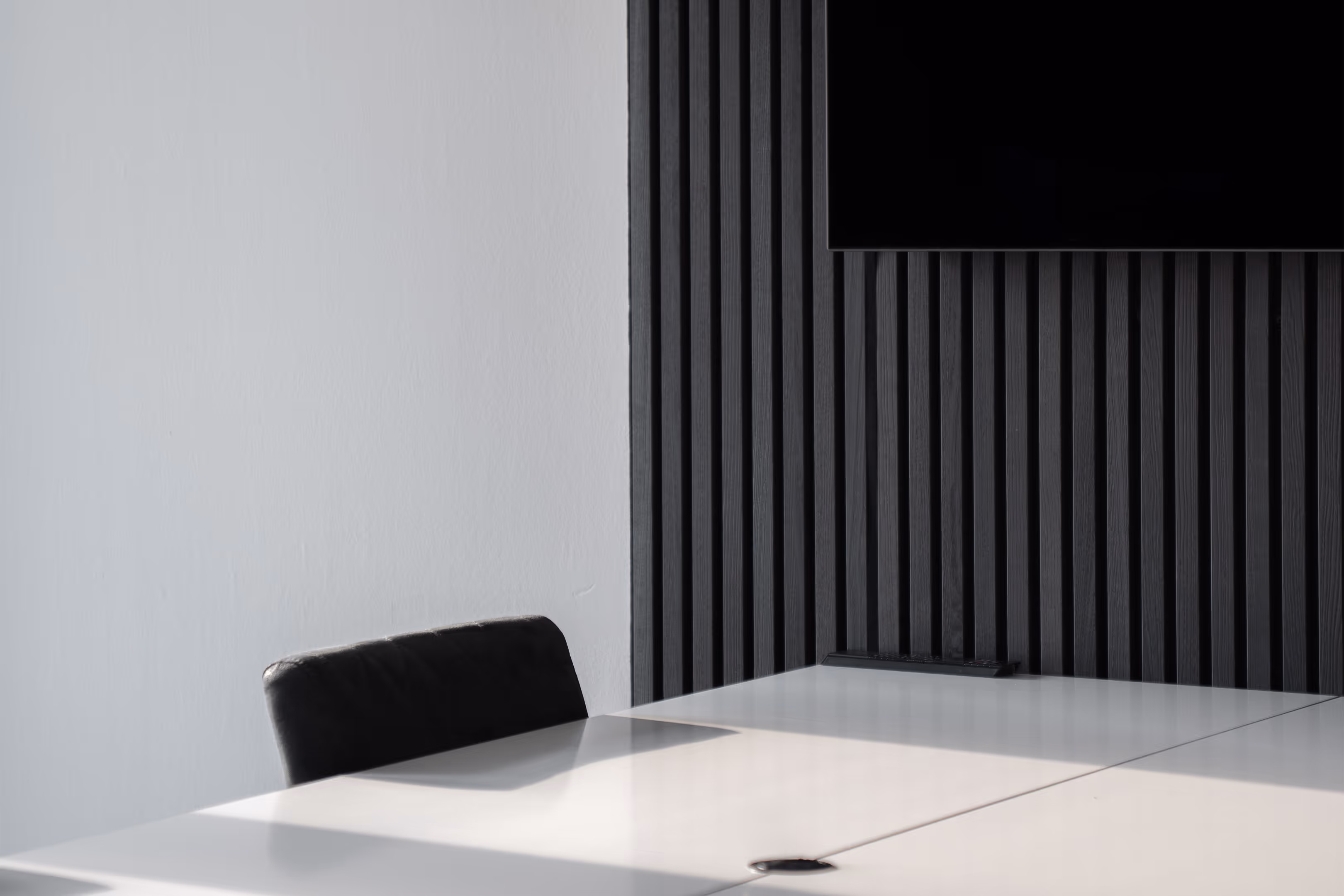 Moderner Konferenztisch mit schwarze Lederstuhl vor weißer Wand und schwarzer vertikaler Holzverkleidung mit Flachbildschirm.