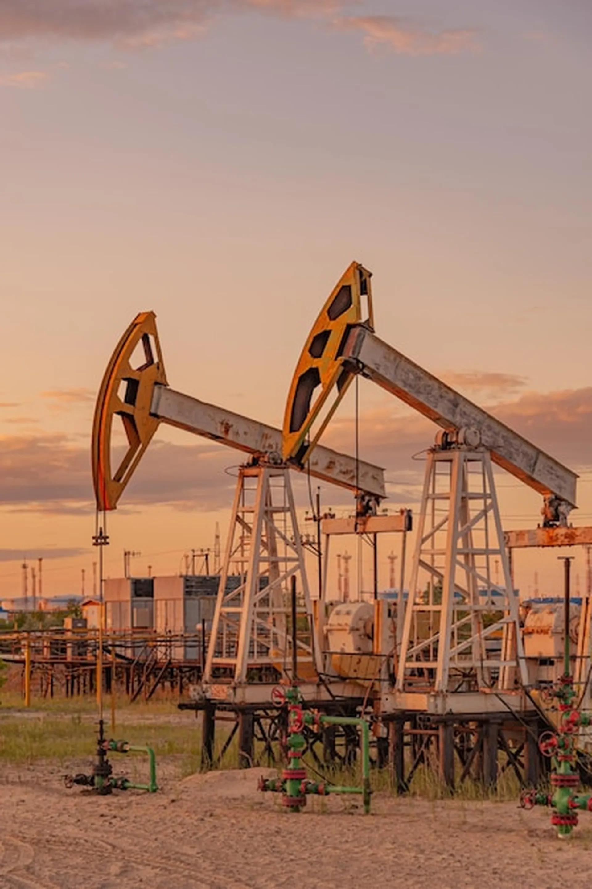 Two oil pump jacks operating in an oil field at sunset with industrial equipment in the background.