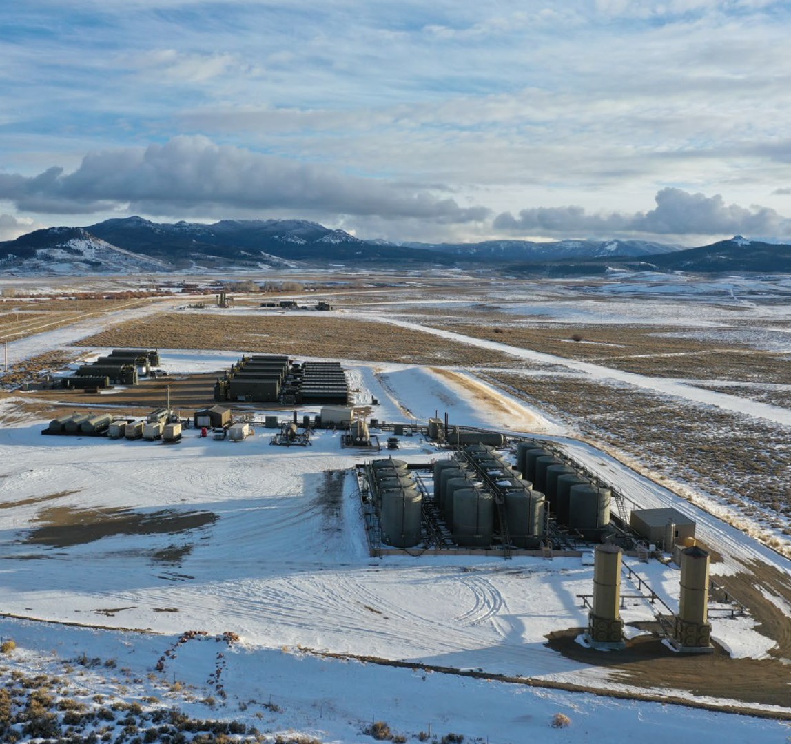 Aerial view of a snowy industrial facility with large storage tanks, equipment, and mountainous landscape in the background.