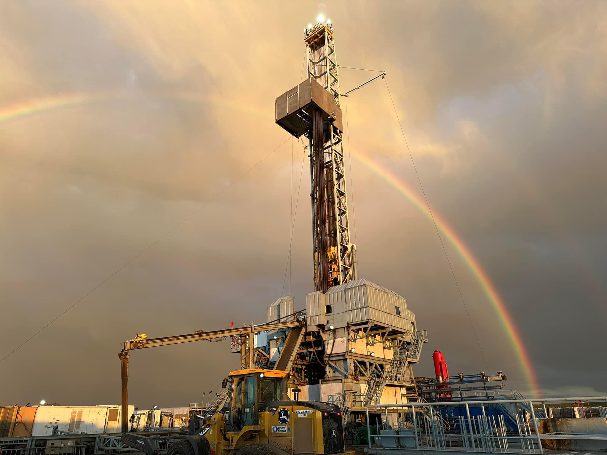 Oil drilling rig and equipment under a cloudy sky with a bright rainbow in the background.