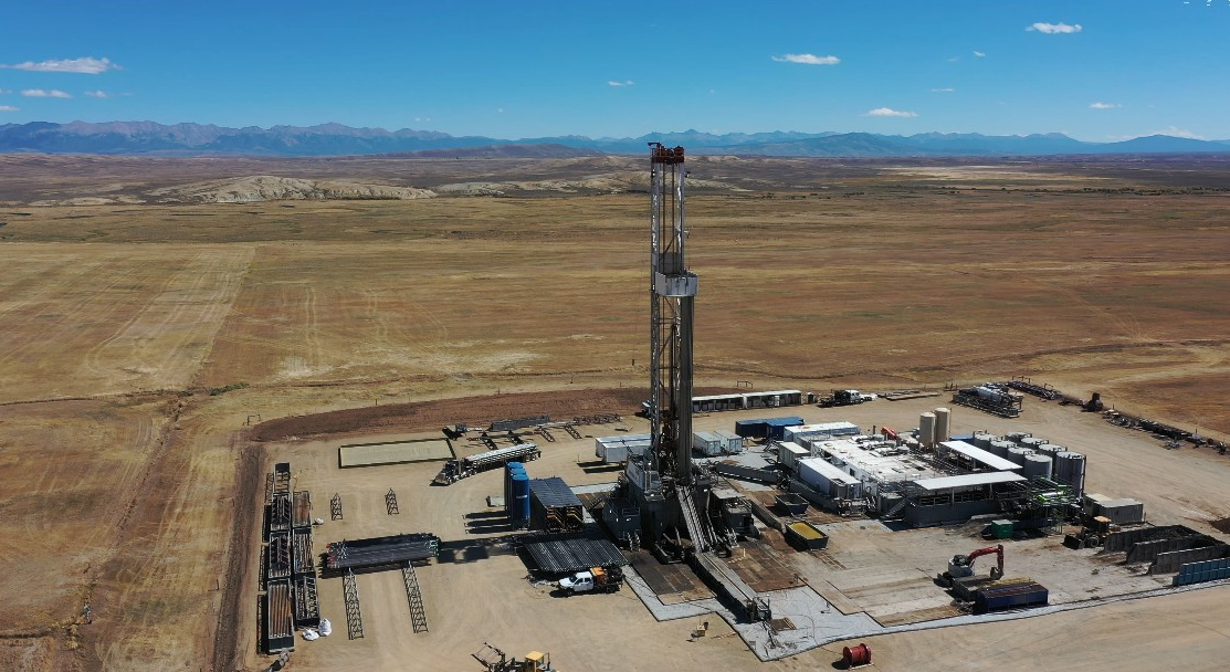 Oil drilling rig with equipment on flat plain, mountains in distance under blue sky with clouds.