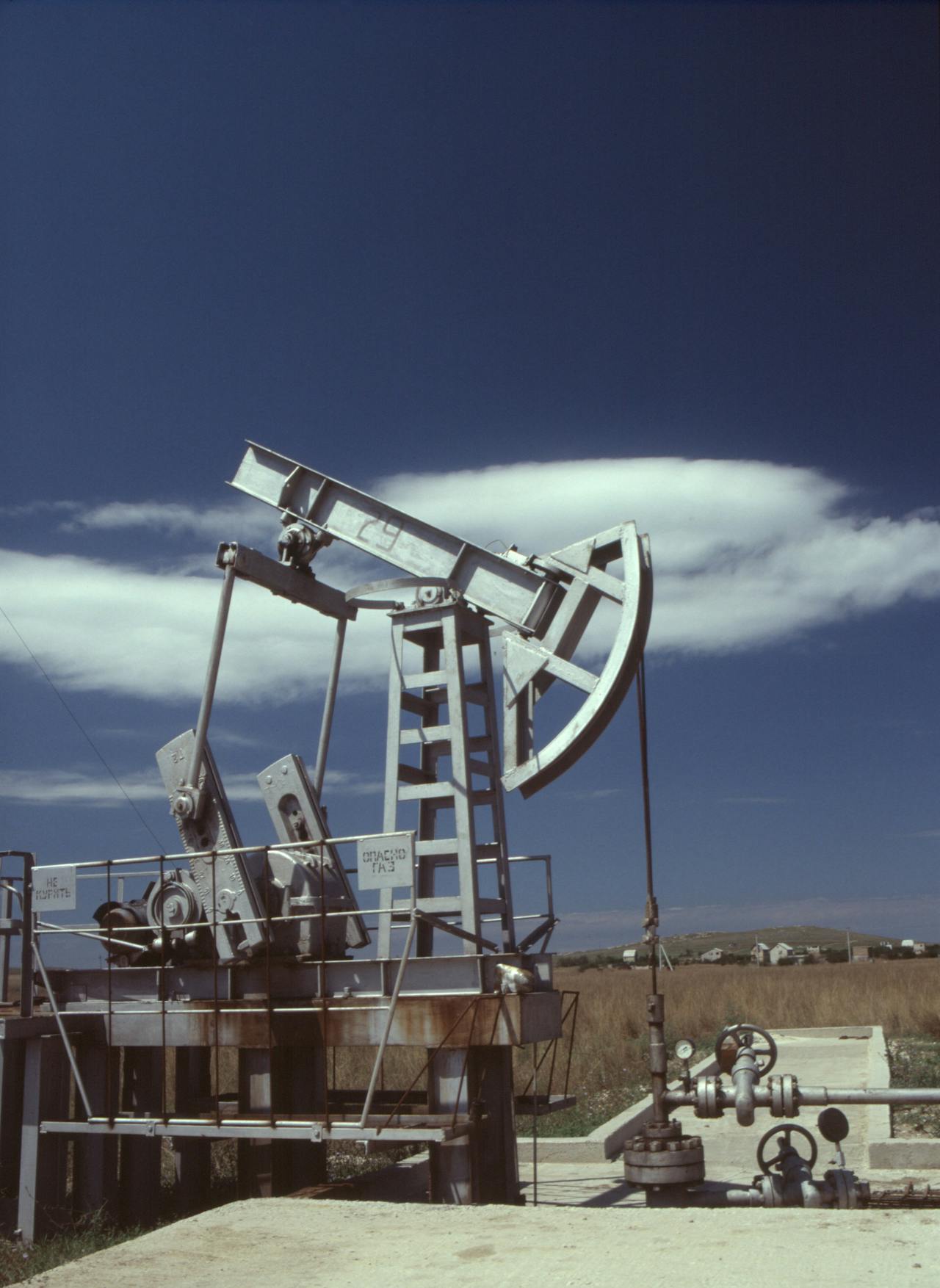 Metal oil pumpjack in a field with a clear sky and distant houses.