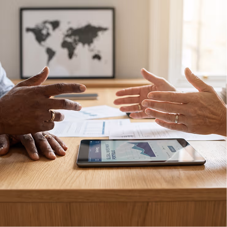 Close-up of two people discussing financial data with a tablet displaying a global investment portfolio graph on a wooden desk.