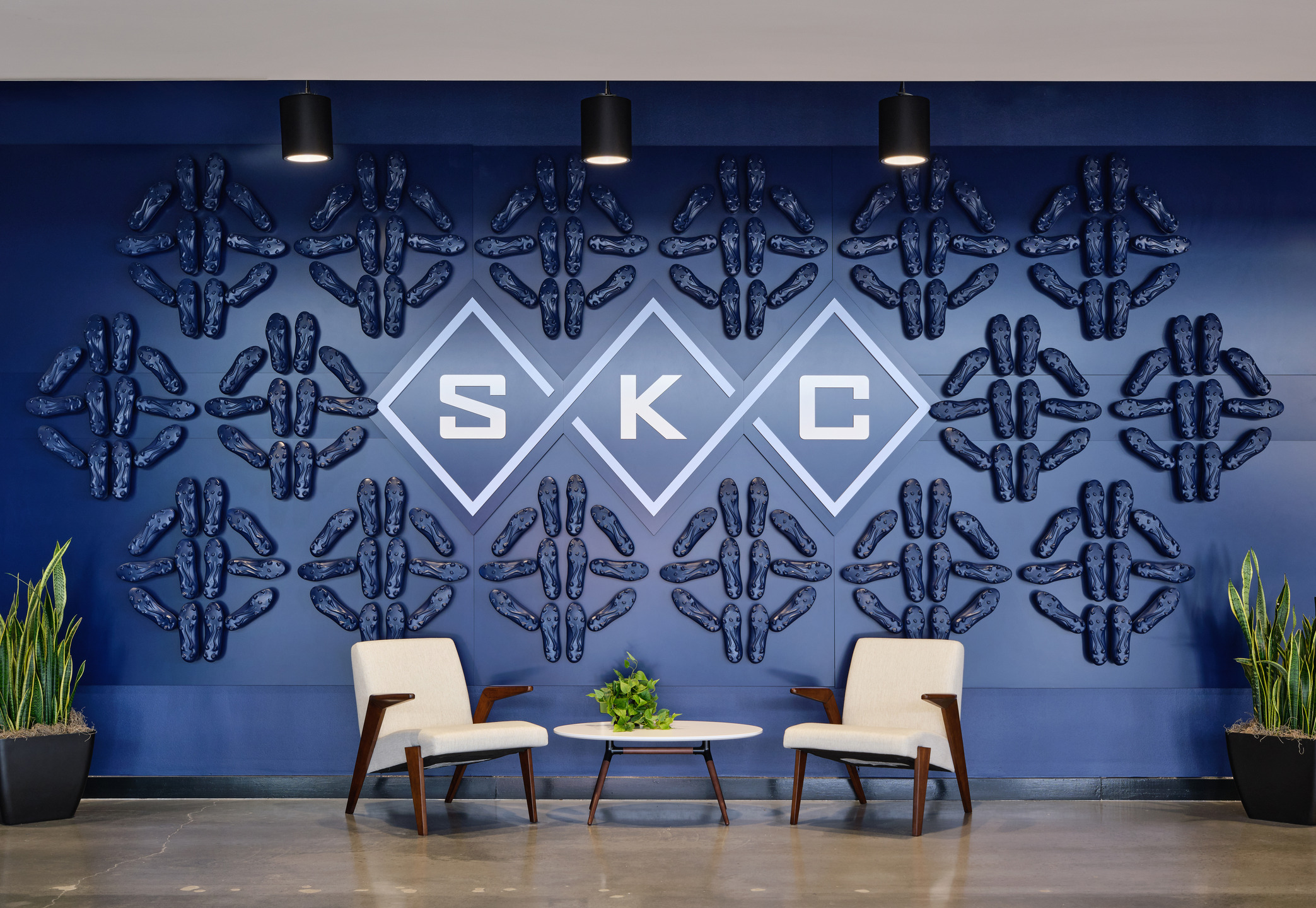 Two white chairs sit in front of a dark blue wall adorned with darker blue soccer cleats arranged in a pattern of diamonds. 