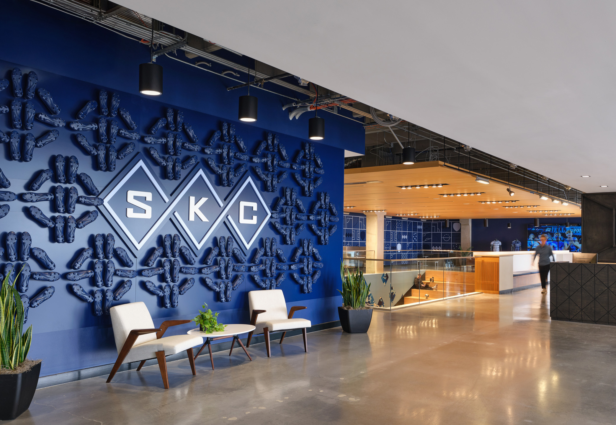 Two white chairs sit in front of a dark blue wall adorned with darker blue soccer cleats arranged in a pattern of diamonds. A light wood grandstand is visible beyond the edge of the blue wall.