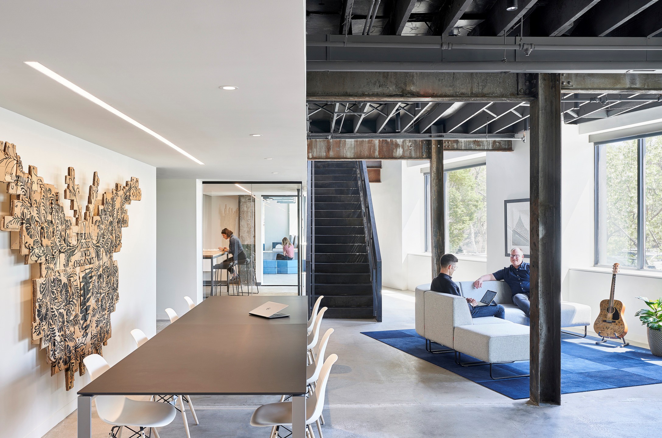 A long table sits to the left of a sofa. Open-air workplace encourage collaboration between different groups and spaces.
