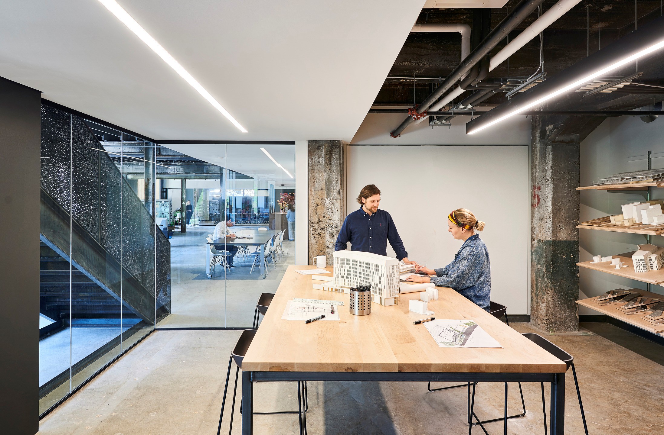 Two people collaborate on a building model at a wood-topped work table in a glass-walled room. 