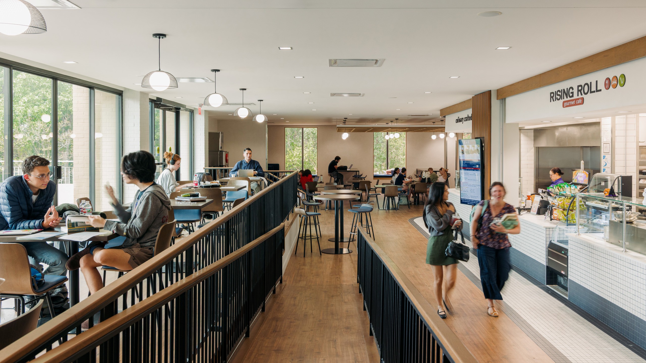 Students sit and stroll through a multi-level dining space, surrounded on two sides with windows.