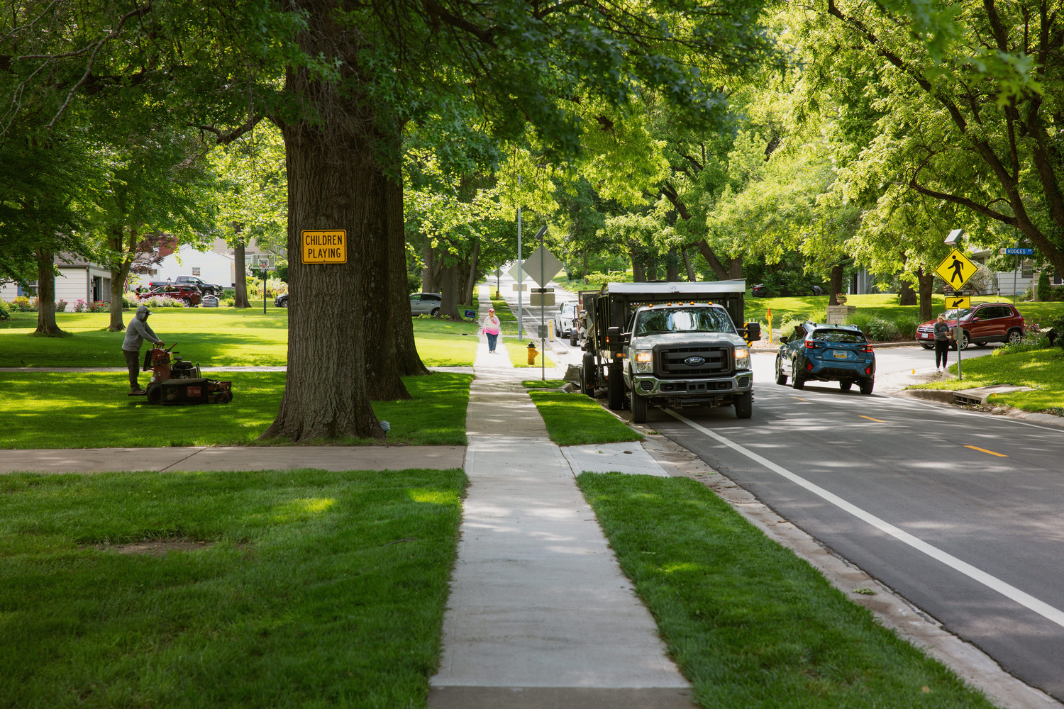 A mower tends to a yard while pedestrians walk along suburban, tree-lined sidewalks.