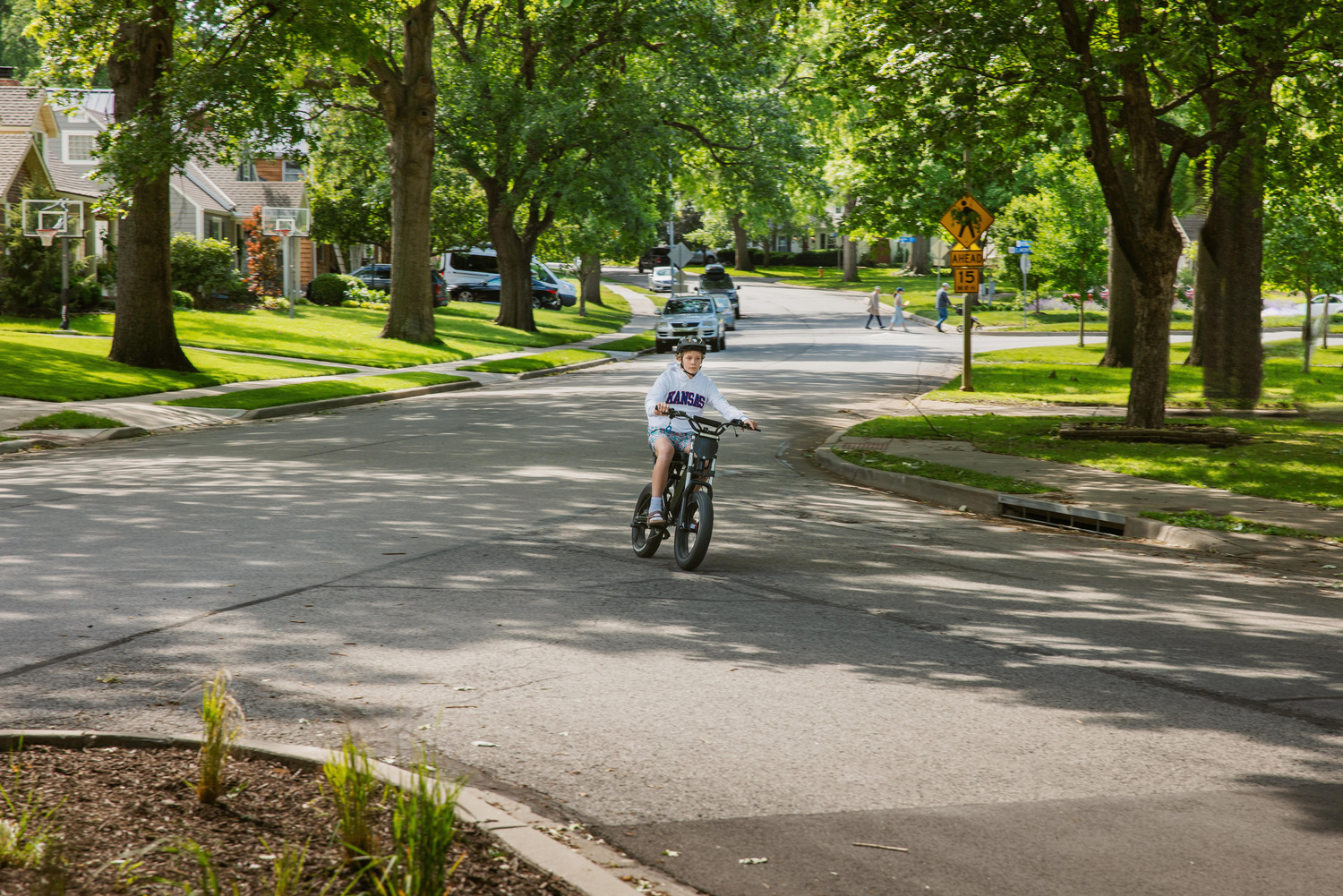 A child rides his bicycles through a suburban, tree-lined street.