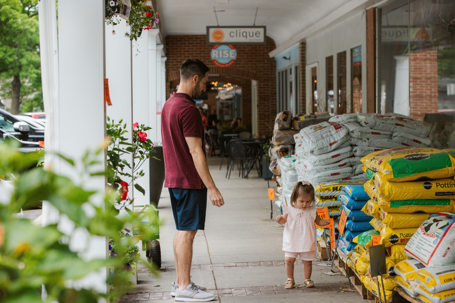 A young child toddles in front of a yard supply store with her father. 