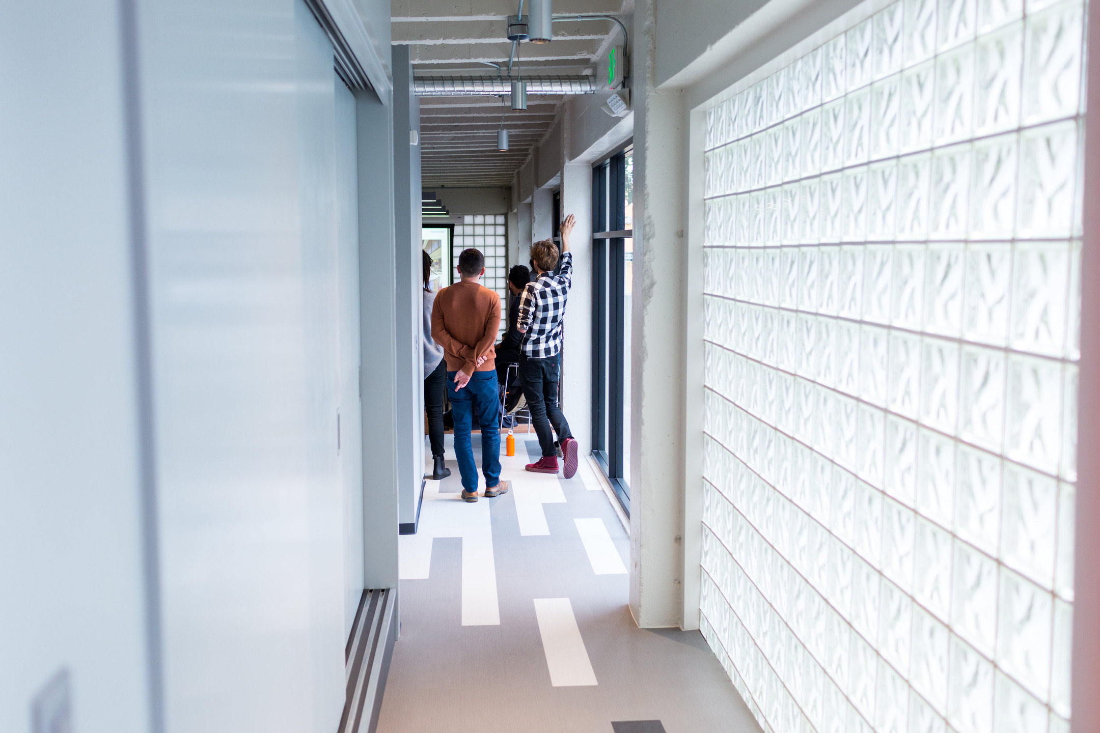 People stand at the end of a long hallway, with one wall constructed from glass tiles.