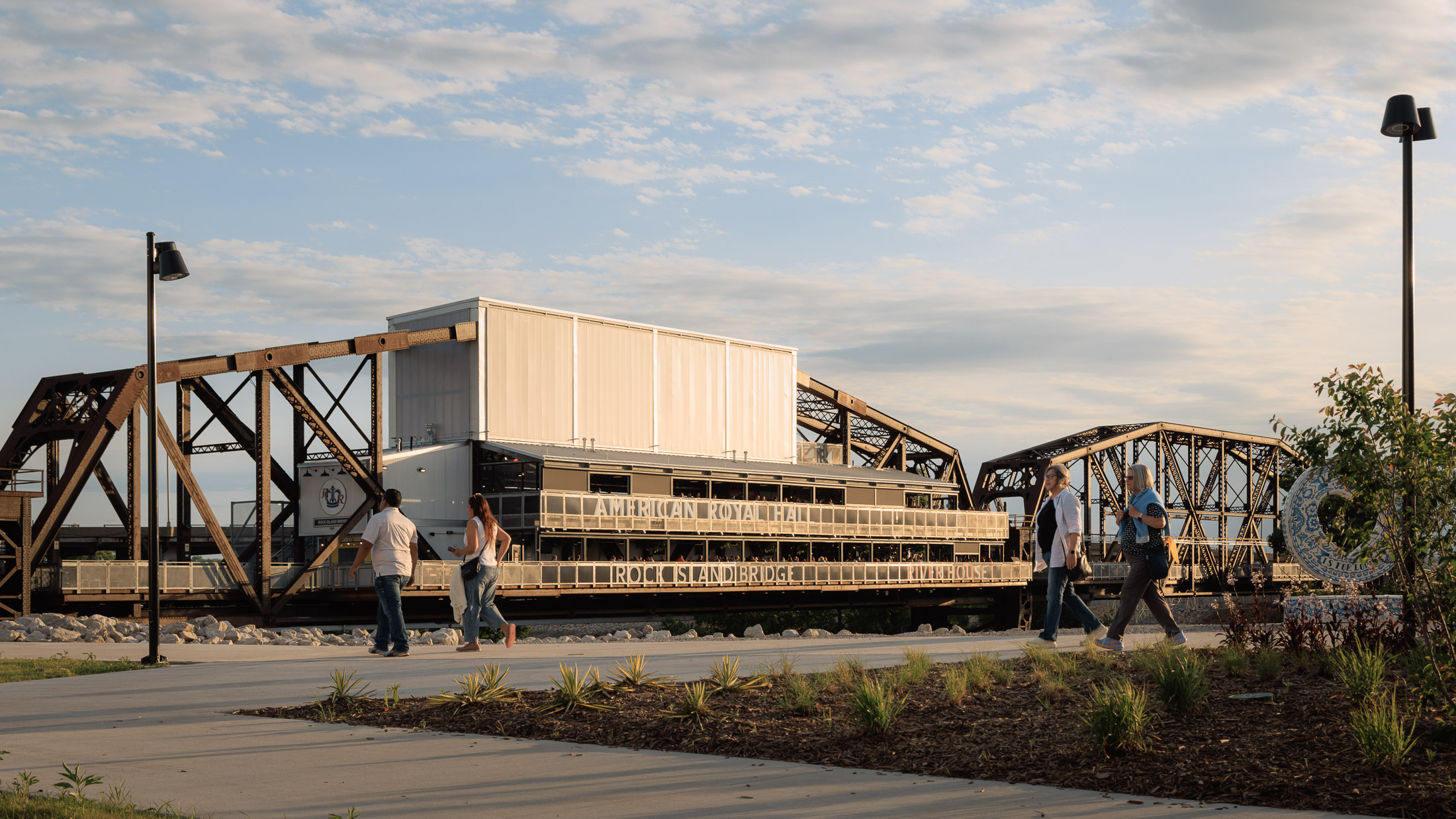 Image of truss bridge spanning tree-lined river with people in the foreground at adjacent park.