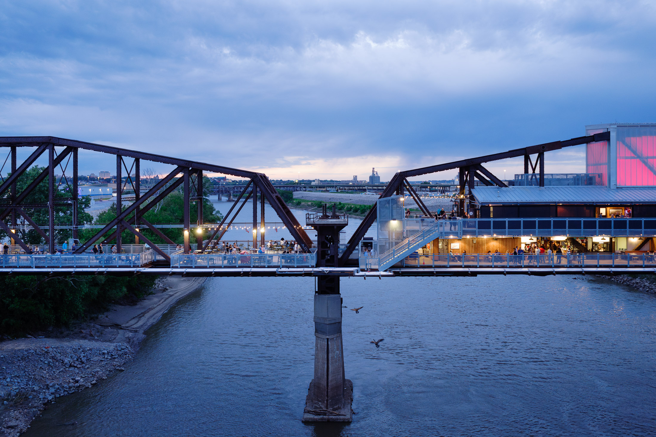 Image of truss bridge spanning river at dusk with people gathering on multiple levels and geese flying by.