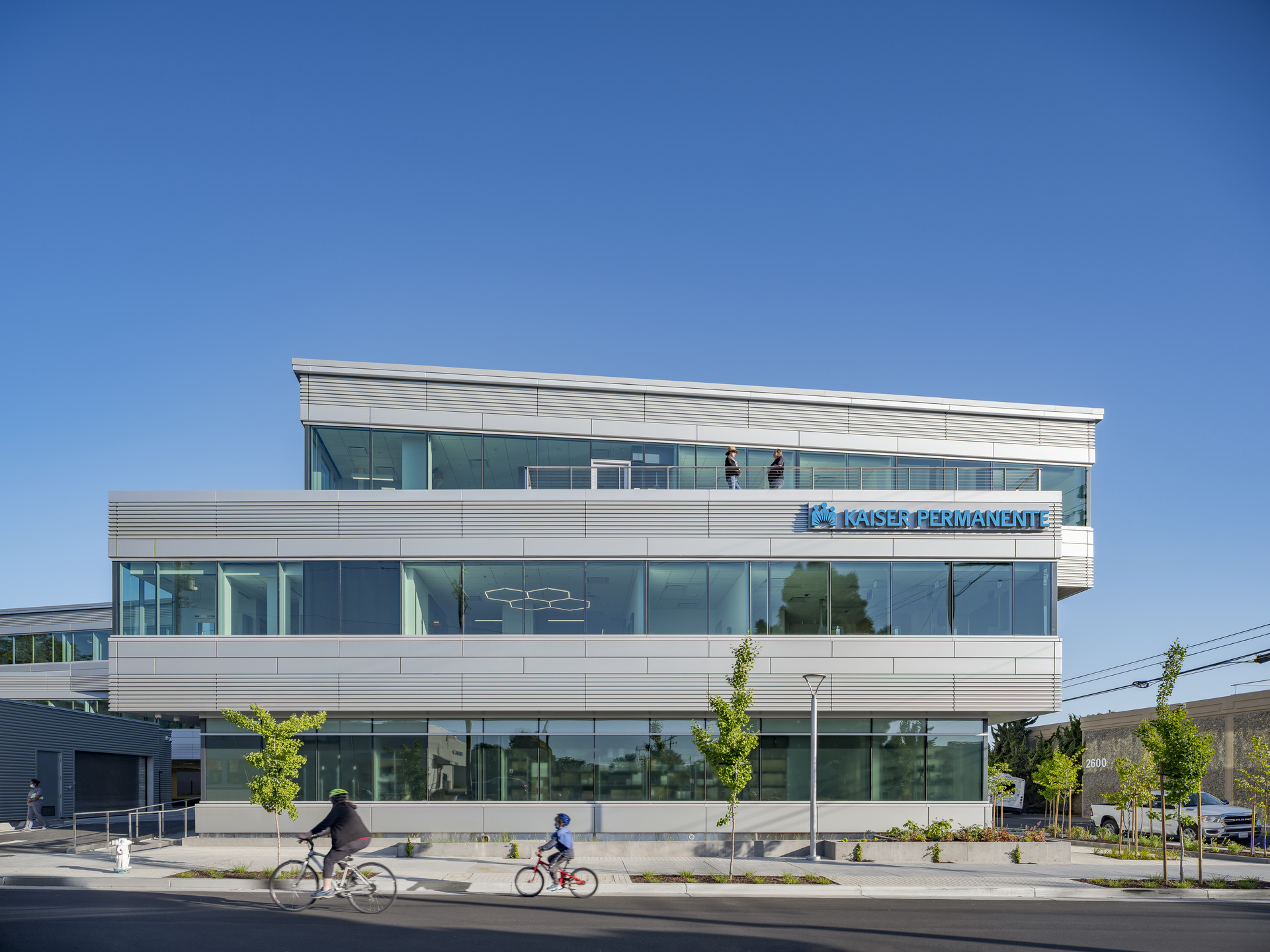 View of the Kaiser Permanente Medical Offices from Parker Street with bicyclists riding by and two people on the rooftop terrace.