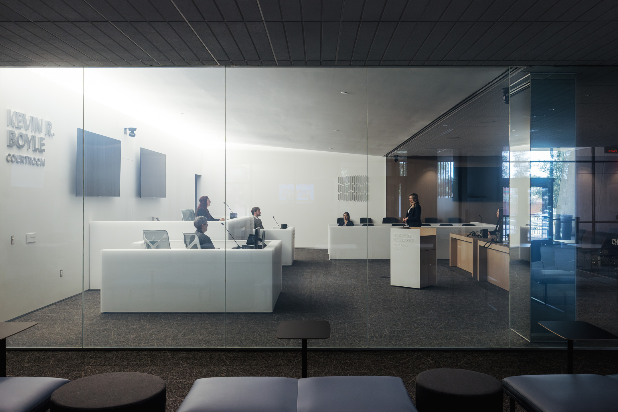 A view through a glass wall of students sit and stand in a mock courtroom, practicing litigation at the University of Arizona College of Law.