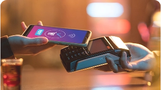 Uma pessoa realiza um pagamento por aproximação (contactless). Um smartphone é segurado próximo a uma maquininha de cartão (POS). Na tela do celular, é visível a palavra "PAY" e o símbolo de pagamento por aproximação. O fundo está desfocado, em tons quentes, sugerindo um ambiente como um bar ou restaurante, com um copo de bebida visível no canto inferior esquerdo.