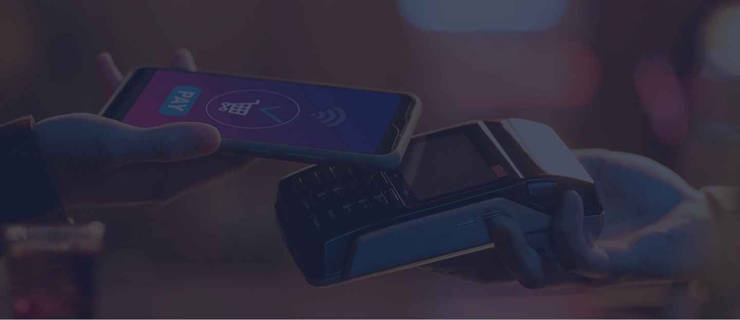 Uma pessoa realiza um pagamento por aproximação (contactless). Um smartphone é segurado próximo a uma maquininha de cartão (POS). Na tela do celular, é visível a palavra "PAY" e o símbolo de pagamento por aproximação. O fundo está desfocado, em tons quentes, sugerindo um ambiente como um bar ou restaurante, com um copo de bebida visível no canto inferior esquerdo.