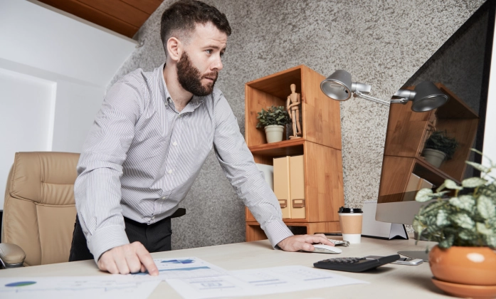 Um homem de barba e camisa social listrada está em pé, inclinado sobre sua mesa de escritório. Ele olha com concentração para a tela do computador, com a mão direita no mouse e a esquerda apontando para um gráfico num documento. No ambiente de trabalho, há também uma calculadora, um copo de café e uma planta. Ao fundo, vê-se uma cadeira de escritório, uma estante de madeira e uma parede cinza com textura.