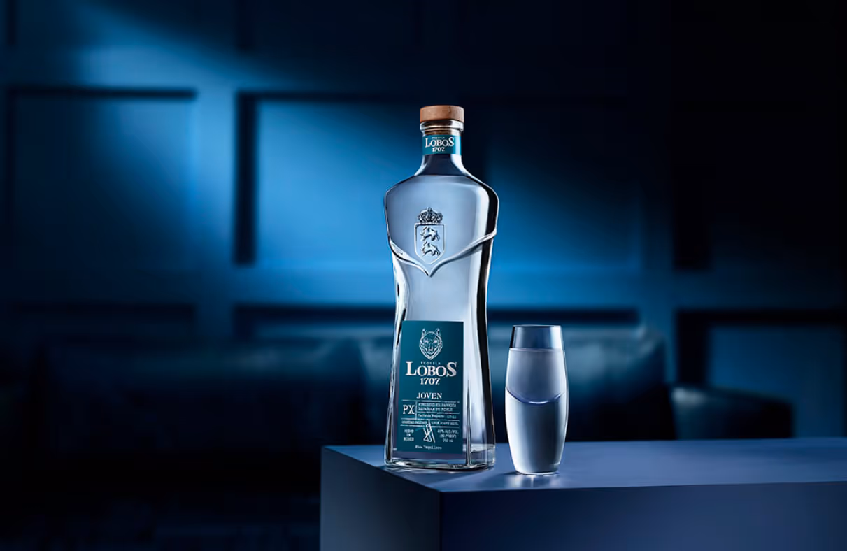 Lobos 1707 Joven Tequila bottle next to a glass of it with a blue background. 