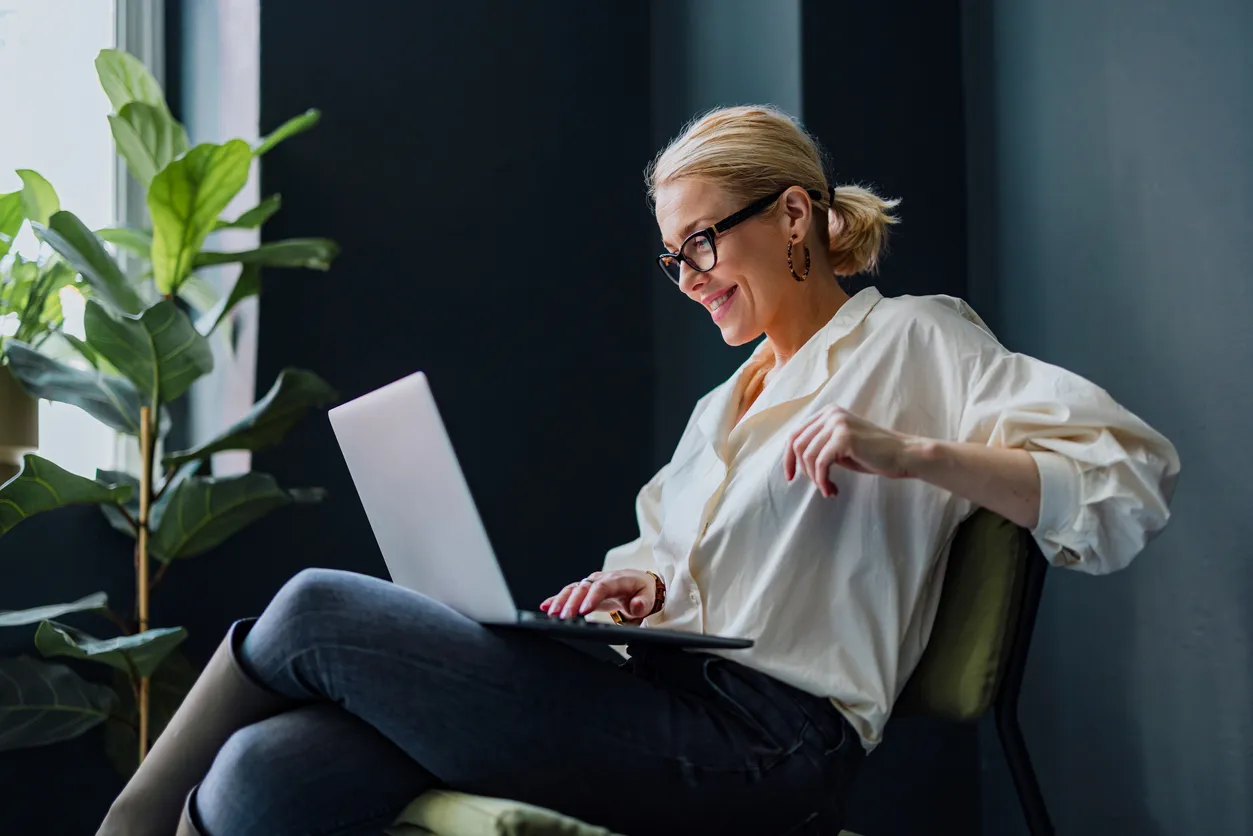 A woman wearing stylish glasses sits comfortably in a modern space, working on a laptop near a large potted plant. This image highlights lifestyle, eye health, and fashion-forward eyewear.
